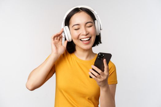 Cute japanese girl in headphones, looking at mobile phone and smiling, using music app on smartphone, standing against white background.