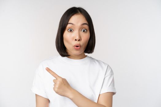 Close up of young japanese woman smiling, pointing finger left at copy space, showing announcement or advertisment banner, standing over white background.