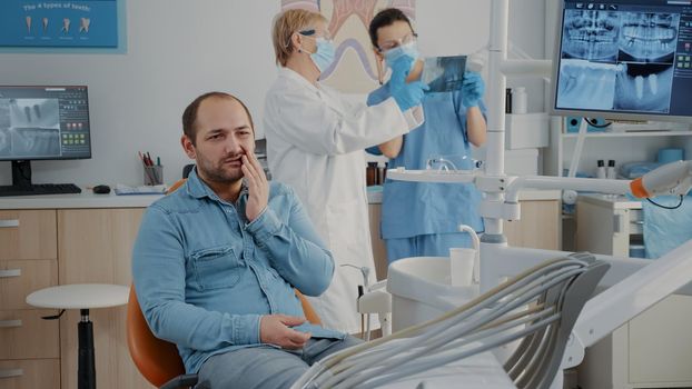 Dentistry patient expressing serious toothache in oral care cabinet. Hurt man dealing with painful caries and denture problems, attending dental appointment to receive treatment.