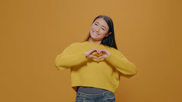 Portrait of woman showing heart shape gesture in studio, doing affectionate love sign to express feelings and romance. Romantic person advertising authentic valentine symbol on camera.