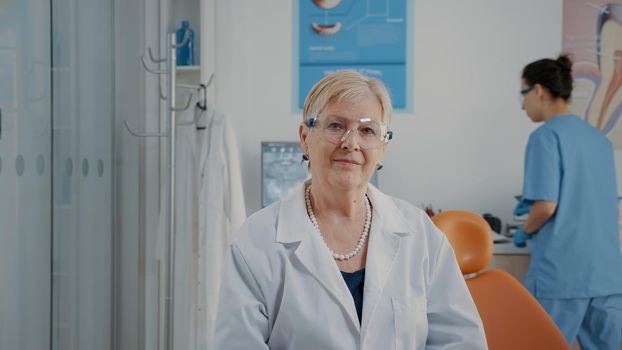 Portrait of mature stomatologist looking at camera in cabinet, working with dental tools for oral care. Woman orthodontist wearing protective glasses to treat dentition problems