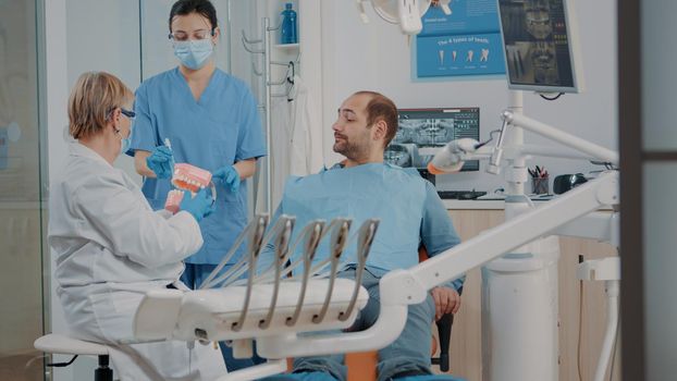 Dentist showing correct way to clean denture with toothbrush, holding artificial jaw for example. Stomatologist giving hygiene lesson to patient learning to brush teeth. Man preventing caries