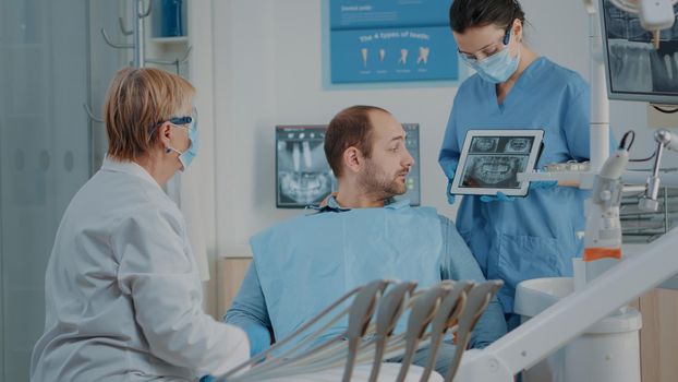 Dentist and nurse showing x ray results to patient with toothache, explaining dental procedure and diagnosis. Stomatology team analyzing radiography to do oral care surgery on man.