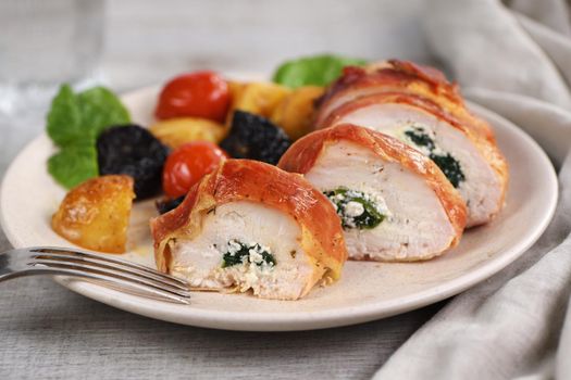 Sliced chicken breast stuffed with goat cheese with spinach, wrapped in prosciutto, with a side dish of baked potatoes, tomato and dried prunes