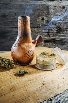 Old brown steamung bong and open cannabis grinder with chopped marijuana weed and medical cannabis bud in smoke by wooden desck and sackcloth. Vertical orientation