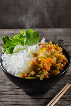 A plate of rice noodles stands on an old wooden table top. Rice noodles are most common in the cuisines of East, Southeast Asia and South Asia. The side dish consists of fried onions, carrots, paprika. Copy space. Vertical orientation