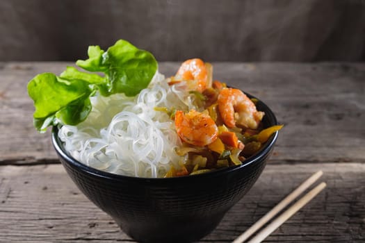 A plate of rice noodles stands on an old wooden table top. Rice noodles are most common in the cuisines of East, Southeast Asia and South Asia. The side dish consists of fried onions, carrots, paprika, shrimp. Copy space. Horizontal orientation