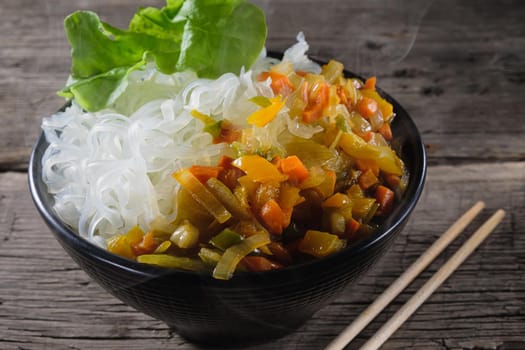 A plate of rice noodles stands on an old wooden table top. Rice noodles are most common in the cuisines of East, Southeast Asia and South Asia. The side dish consists of fried onions, carrots, paprika. Copy space. Horizontal orientation