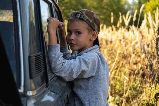 a cute six year old boy stands by the car against the background of autumn grass.. horizontal orientation.
