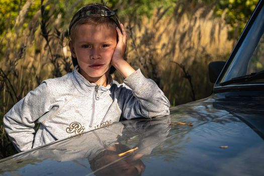 a cute six year old boy stands by the car against the background of autumn grass.. horizontal orientation.