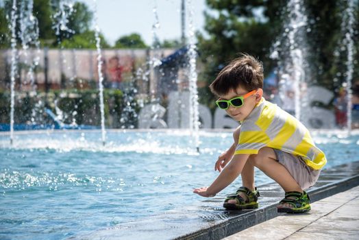 Little boy plays in the square near pool with water jets in the fountain at sunny summer day. Active summer leisure for kids in the city.