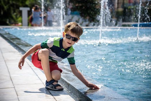 Little boy plays in the square near pool with water jets in the fountain at sunny summer day. Active summer leisure for kids in the city.