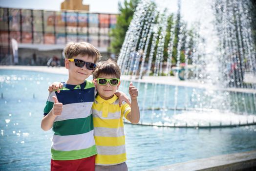 Group of happy children playing outdoors near pool or fountain. Kids embrace show thumb up in park during summer vacation. Dressed in colorful t-shirts and shorts with sunglasses. Summer holiday concept.