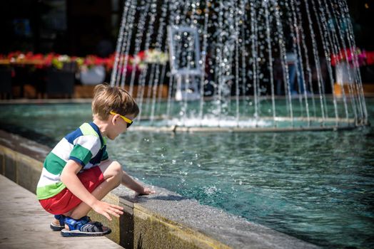 Little boy plays in the square near pool with water jets in the fountain at sunny summer day. Active summer leisure for kids in the city.