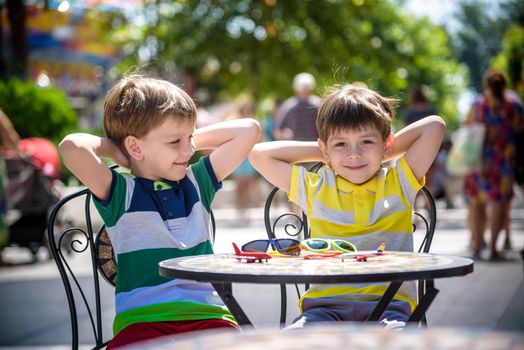 Two little kid boys waiting on table for healthy breakfast in hotel restaurant or city cafe. Child sit on comfortable chair with hands up, relaxed, enjoy his vacation. Summer holiday with children concept.