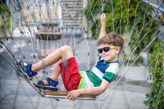 Little adorable kid sitting on chain swing during summer holidays on vacation. Boy relaxed and happy wearing casual clothes in sunglasses. Active summer leisure for kids in the city concept.