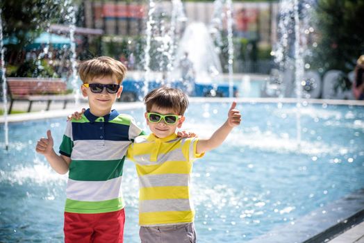 Group of happy children playing outdoors near pool or fountain. Kids embrace show thumb up in park during summer vacation. Dressed in colorful t-shirts and shorts with sunglasses. Summer holiday concept.