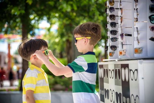 A child and a choice of sunglasses. Two little boys are standing in sun-proof glasses against the background of a shop window with glasses. Kid help choose each other. Sunglasses sale during summer vocation.