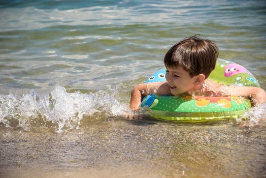 Happy little boy playing with colorful inflatable ring in outdoor swimming sear river lake or pool on hot summer day. Kids learn to swim and having fun.