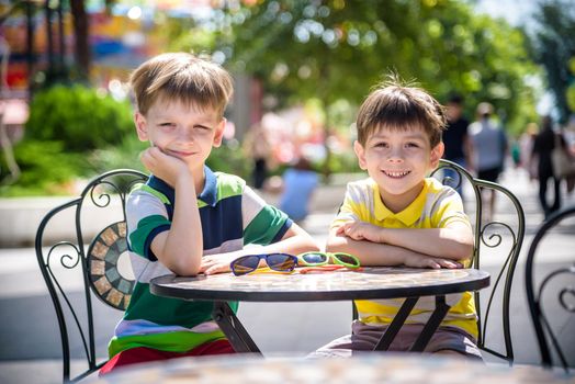 Two little kid boys waiting on table for healthy breakfast in hotel restaurant or city cafe. Child sit on comfortable chair, relaxed, enjoy his vacation. Summer holiday with children concept.