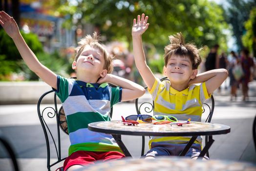 Two little kid boys waiting on table for healthy breakfast in hotel restaurant or city cafe. Child sit on comfortable chair with hands up, relaxed, enjoy his vacation. Summer holiday with children concept.