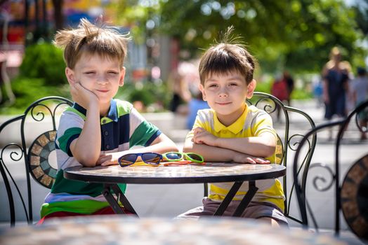 Two little kid boys waiting on table for healthy breakfast in hotel restaurant or city cafe. Child sit on comfortable chair, relaxed, enjoy his vacation. Summer holiday with children concept.