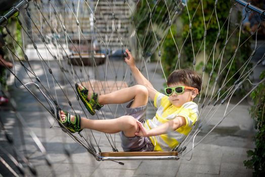 Little adorable kid sitting on chain swing during summer holidays on vacation. Boy relaxed and happy wearing casual clothes in sunglasses. Active summer leisure for kids in the city concept.