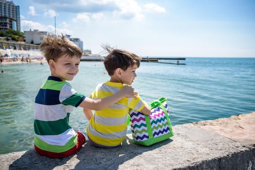 Two beautiful little boy sibling brothers in vest and shorts sitting on breakwater against the sea and shore in summer. Preschool children best friends enjoying summer vacations on sea. View from back.