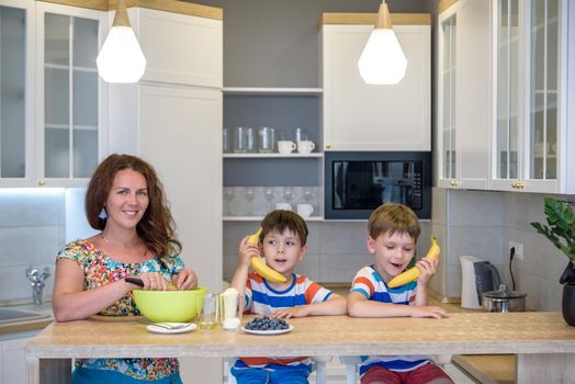 Cute little sibling brother boys cook playing with a banana, as if it were a phone. Mother prepare dough on modern kitchen.