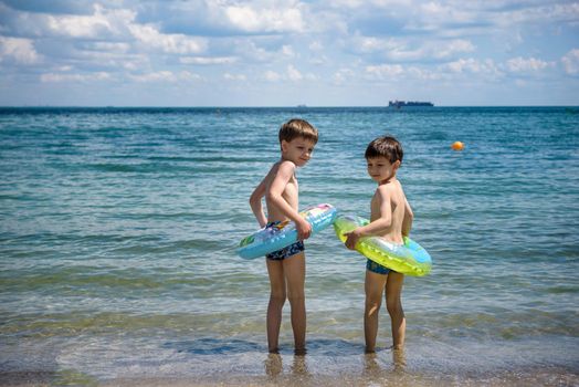 Two cute little brother boy stand on the beach in swimming trunks goes swimming with inflatable ring. Safe water activities with kids.