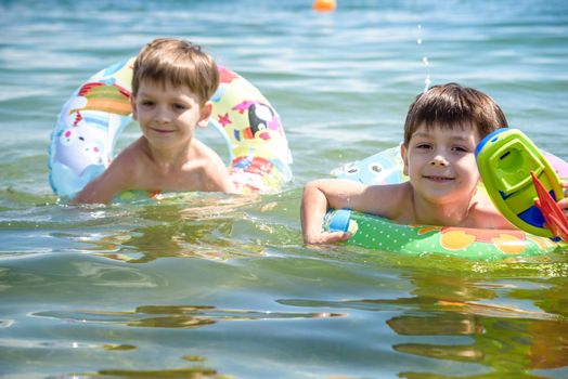Happy kids have fun in sea surf on beach. Joyful couple of children on inflatable ring ride on breaking wave. Travel lifestyle, swimming activities in family summer camp. Vacations on tropical island.