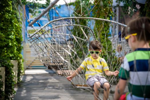 Little adorable kid sitting on chain swing during summer holidays on vacation. Boy relaxed and happy wearing casual clothes in sunglasses. Active summer leisure for kids in the city concept.