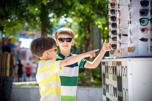A child and a choice of sunglasses. Two little boys are standing in sun-proof glasses against the background of a shop window with glasses. Kid help choose each other. Sunglasses sale during summer vocation.