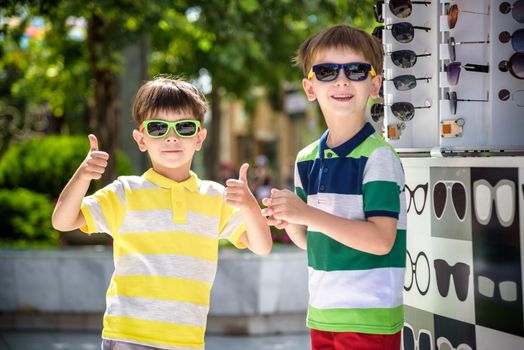 A child and a choice of sunglasses. Two little boys are standing in sun-proof glasses against the background of a shop window with glasses. Kid help choose each other. Sunglasses sale during summer vocation.