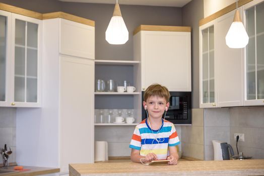 Little boy sitting at kitchen table holding a phone in his hands with earphones while looking to screen, messaging online using Wi-Fi having astonished expression.