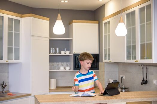 Smart preteen schoolboy doing his homework with digital tablet at home. Child using gadgets on his kitchen to study. Modern education and learning for kids.