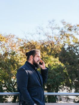 Young businessman with long hair and beard calling during his work break, on the street next to the offices. Vertical photo on a sunny and clear day. Size view