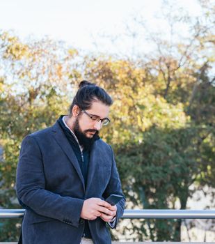 Young businessman with long hair and beard looking smartphone during his work break, on the street next to the offices with glasses. Vertical photo on a sunny and clear day.