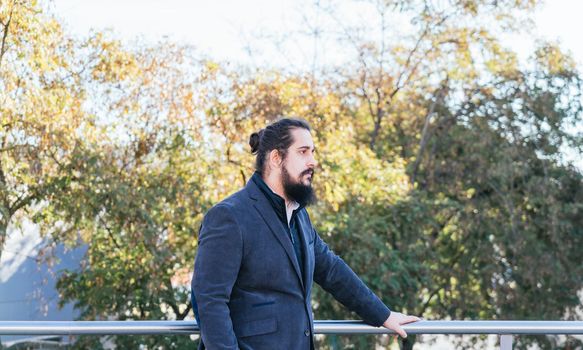 Young businessman with long hair and beard standing minding his own business during his work break, on the street next to the offices. Horizontal photo on a sunny and clear day.