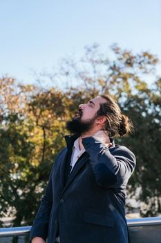 Young businessman with neck pain from work. He massages his neck near his offices on the street. Young entrepreneur with long hair and beard, brown, on a sunny day.