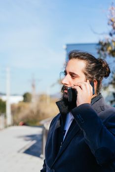 Young businessman with long hair and beard calling during his work break, on the street next to the offices. Vertical photo on a sunny and clear day.