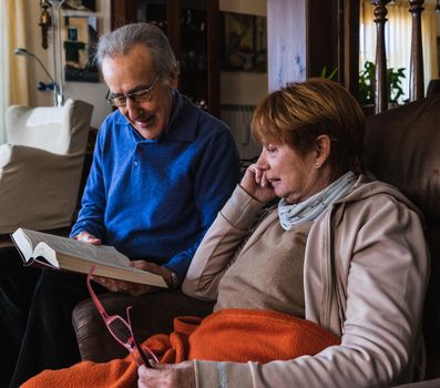 Grandparents reading together a book in living room. He have a blue sweater and is supporting the book. She is sited on the sofa reading with purple glasses in a hand