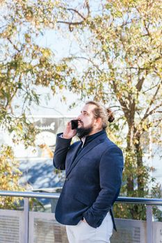 Young businessman with long hair and beard calling during his work break, on the street next to the offices. Vertical photo on a sunny and clear day.