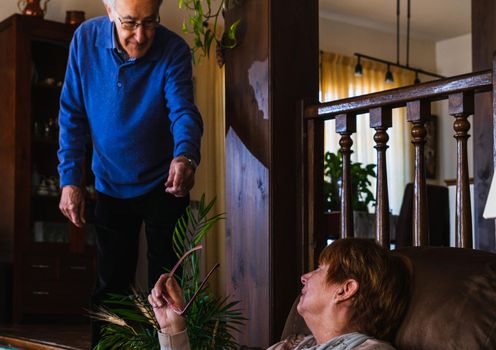 Old man saying something while stand up in living room to his wife. She is sited on a sofa with purple glasses in hand. They are caucasian old couple. They are in living room with natural light