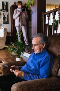 Grandfather using tablet in a living room with natural light. He wear a blue sweater and glasses. Its in his house, size view with natural light