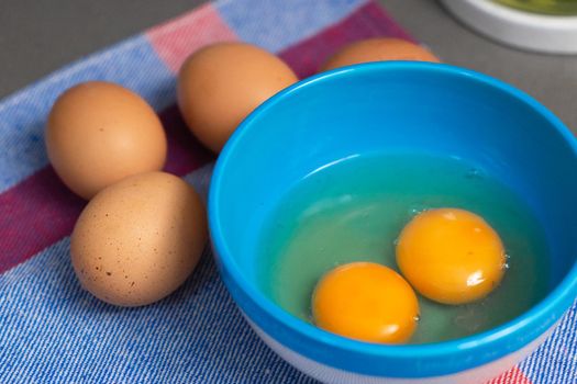 two egg yolks in a blue bowl for a festive sweet or savoury recipe. the blue bowl is placed behind a raw natural eggs up to a blue cloth. In the upper right edge are a oil mill. There are all on a gray countertop