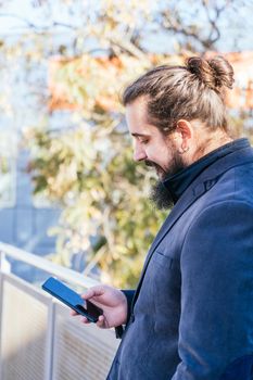 Young businessman with long hair and beard looking smartphone during his work break, on the street next to the offices. Vertical photo on a sunny and clear day. Size view.