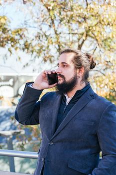 Young businessman with long hair and beard calling during his work break, on the street next to the offices. Vertical photo on a sunny and clear day.