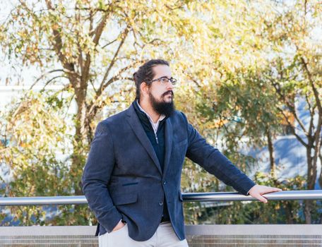 Young businessman with long hair and beard standing minding his own business during his work break, on the street next to the offices with glasses. Horizontal photo on a sunny and clear day.