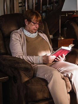Old woman reading a reed book with house clothes in a sofa. Grandmother reading in house with natural light.